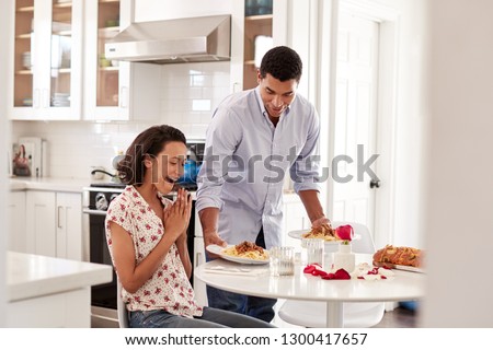 Young African American adult woman sitting at the table in the kitchen, her partner surprising her by serving a romantic meal, selective focus