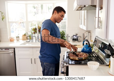 Young adult African American man standing in the kitchen cooking on the hob, using a spatula and frying pan, side view, close up