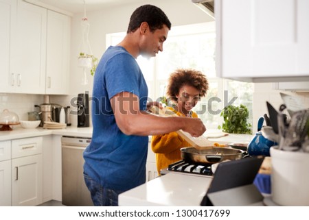 African American pre-teen girl and her father standing at hob in the kitchen preparing food in a frying pan, following a recipe on a tablet computer, backlit