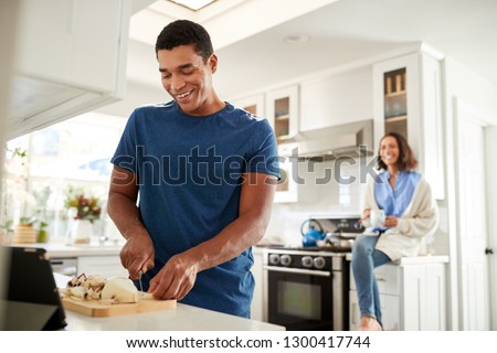 Young African American adult man standing in the kitchen preparing food, his partner sitting on kitchen worktop behind him, focus on foreground