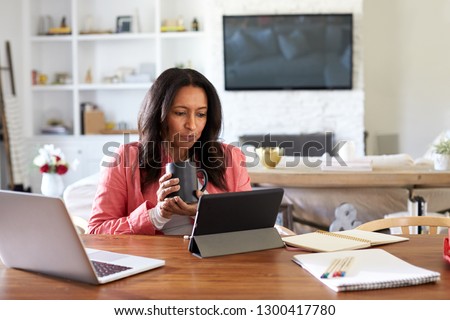 Middle aged woman sitting at a table reading using a tablet computer, holding a cup, front view