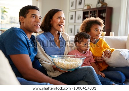 Young African American family sitting together on the sofa in their living room watching TV and eating popcorn, side view