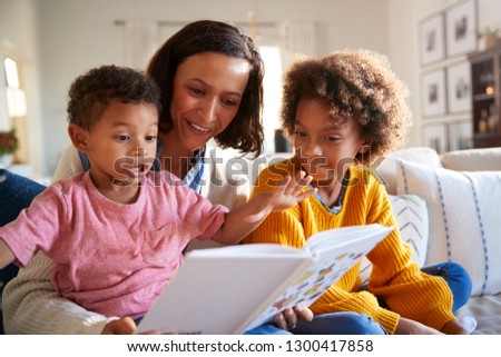 Excited African American toddler boy sitting on his mothers knee reading a book in the living room with his older sister, close up