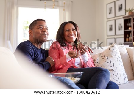 Close up of middle aged African American couple sitting on the sofa in their living room watching TV, laughing and eating popcorn, close up
