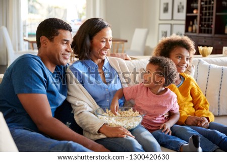 African American toddler boy eating popcorn, sitting on the sofa with his sister and parents in their living room watching a movie together