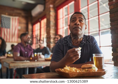 Man Watching Game On Screen In Sports Bar Eating Burger And Fries