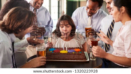 Businesswoman Blowing Out Candles On Birthday Cake At Celebration In Bar With Colleagues
