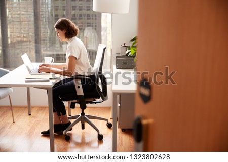 Businesswoman Sitting At Desk Working On Laptop In Modern Office Viewed Through Door Way