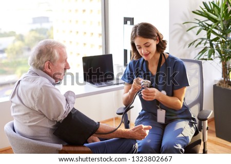 Nurse Wearing Scrubs In Office Checking Senior Male Patients Blood Pressure