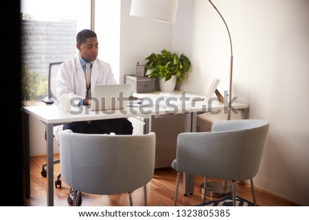 Male Doctor Wearing White Coat In Office Sitting At Desk Working On Laptop