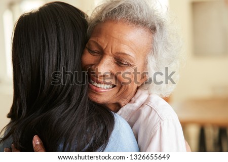 Over The Shoulder View Of Senior Mother Being Hugged By Adult Daughter At Home