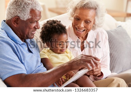 Grandparents Sitting On Sofa At Home Reading Book With Baby Granddaughter