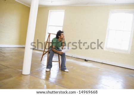 Man sitting on ladder in empty space holding paper thinking