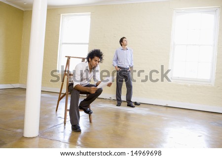 Man sitting on ladder in empty space holding paper with other man behind him