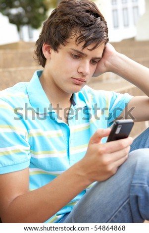Unhappy Male Teenage Student Sitting Outside On College Steps Using Mobile Phone