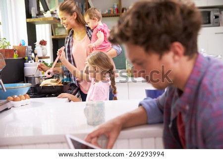 Mother Cooks Family Meal Whilst Father Uses Digital Tablet