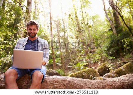 Man Sits On Tree Trunk In Forest Using Laptop Computer