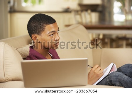 Teenage Boy Sitting On Sofa At Home Doing Homework Using Laptop Computer Whilst Watching TV
