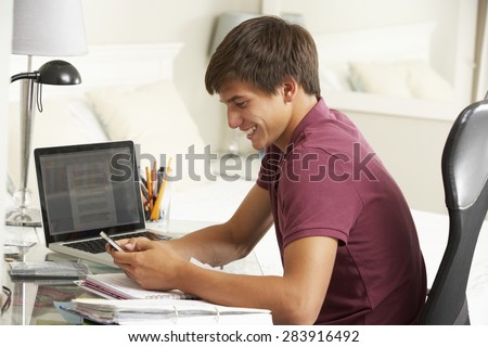 Teenage Boy Studying At Desk In Bedroom Using Mobile Phone