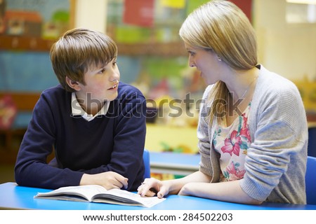 Teacher With Male Pupil Reading At Desk In Classroom