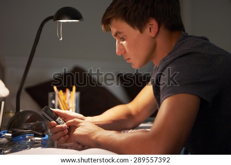 Teenage Boy Texting On Phone Whilst Studying At Desk In Bedroom In Evening
