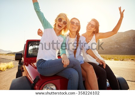 Three Female Friends On Road Trip Sit On Car Hood