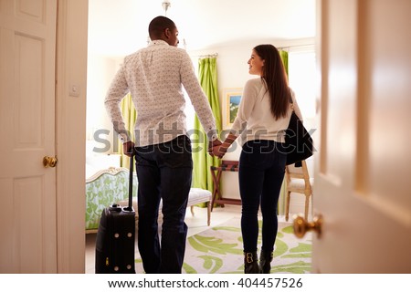 Young multi ethnic couple walk in to a hotel room, back view