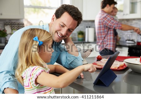 Girl uses tablet in kitchen with dad, while other dad cooks