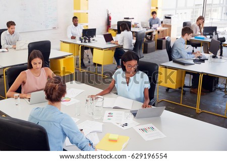 Three women share a desk in a busy office, elevated view
