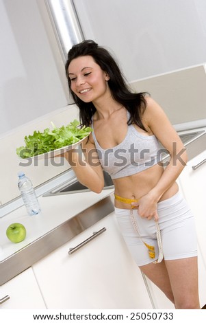 young caucasian brunette in kitchen with a sald dish and a centimeter to measure her hips.Concept of correct food to have a slim body