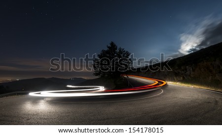 Lights on the asphalt, at night on a mountain road, illuminated by the moon begins to rise over the horizon, a car making a turn, leaving behind a trail of light.