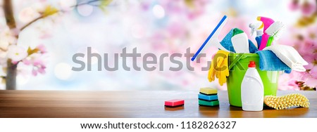 Bucket or basket with cleaning items on wooden table and blurry pink flowers tree background. Washing set with copy space banner.
