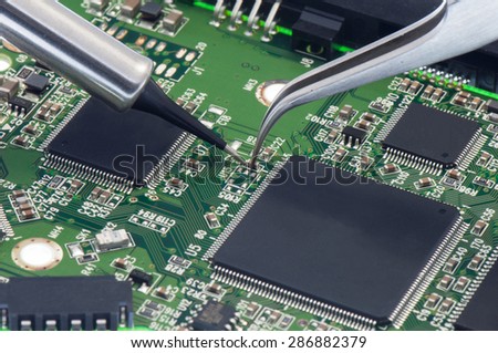 Electronic component  held with tweezers by an engineer over a green motherboard.