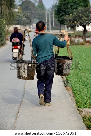 PENGZHOU, CHINA:  Farmer walking down a country road carrying two pails of water suspended from a shoulder yoke