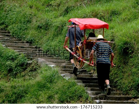 LONGSHENG, CHINA - MAY 2, 2006:  Porters carry a tourist seated in a covered sedan chair up a steep flight of stone stairs to the ancient Yao village of Ping-An in China\'s Guang Xi Province