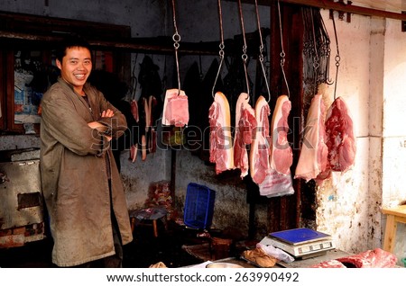 Pengzhou, China - November 20, 2013:  Butcher standing in his shop with various cuts of pork hanging from iron meat hooks
