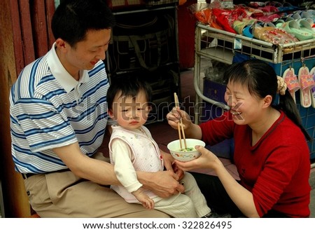 Dujiangyan, China - April 24, 2005:   Mother feeding her toddler son who sits on his father\'s lap in front of their shop