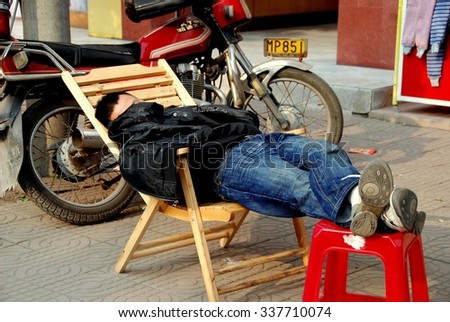 Jiu Chi Town, China - December 1, 2007:  Man uses a wooden chair to stretch out for a nap on a sunny afternoon in front of a clothing shop