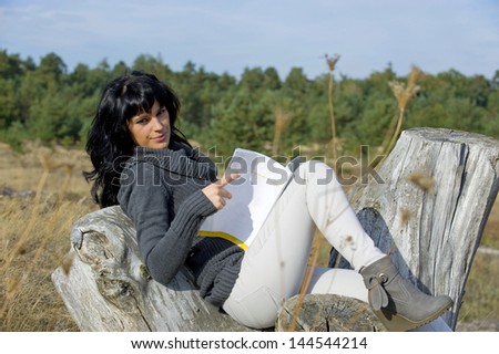 Beautiful and happy young women sitting outside in fall and is reading a book. Image taken on a sunny day. The girl is sitting in front of the forest with the grass on field almost seasonal dry.