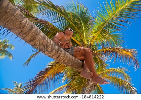 Young beautiful man sitting on the palm tree with coconut cocktail. Having summer fun during travel holidays vacation.