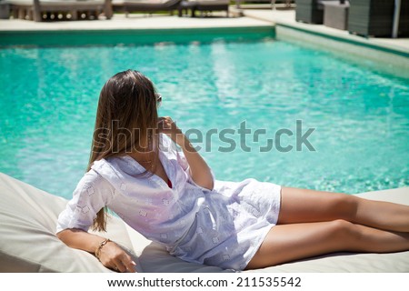 Long haired young woman in white dress lying down on white luxury sofa in pool side, spending summer holidays on tropical resort