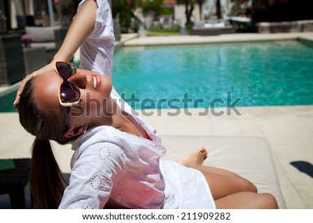 Portrait of sexy cheerful woman holding hand behind head, relaxing at the luxury poolside. Girl at travel spa resort pool. Summer luxury vacation. (focus on woman face)