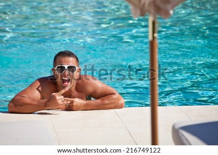 Handsome and happy man relaxing resting on his hands at the side of a sun bathed swimming pool smiling. Summer luxury vacation.