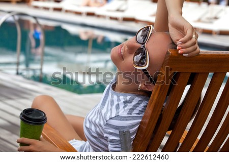 Portrait of cheerful woman holding hand behind head, relaxing at the luxury poolside. Girl at travel spa resort pool. Summer luxury vacation. (focus on woman face)