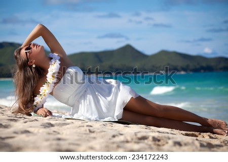 Gorgeous young woman with welcoming Lei in white summer dress laying on the Hawaiian beach. Luxuriating in the sunshine.