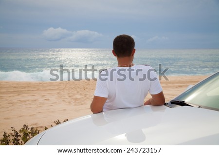 Rear view of young Man leaning on his car parked in front of ocean. Man on road trip enjoying peace and silence relaxing on nature, daydreaming. Vacations And Tourism Concept. Tropical Resort.