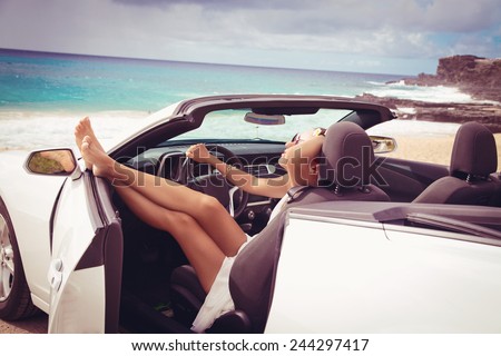 Happy beautiful young woman sitting in a sports car on beautiful sunny summer day. Sexy woman\'s legs showing out of the car, enjoying freedom feeling happy on the Hawaiian beach.