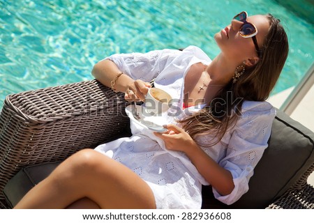 Woman relaxing at the poolside with morning coffee, sunny day, outdoor. Girl at travel spa resort pool. Summer luxury vacation. (focus on woman face)