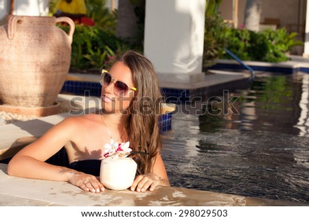 Cute young woman in sexy bikini at edge of a swimming pool with exotic cocktail, coconut. Girl at travel spa resort pool. Summer luxury vacation.