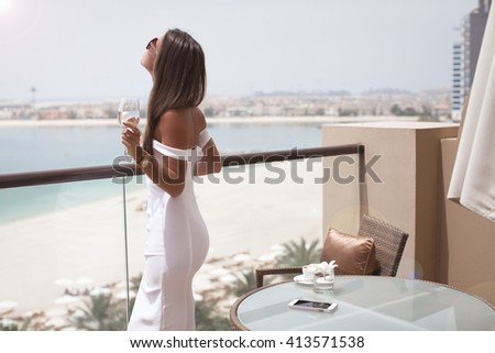 Luxury travel vacation woman with champagne glass looking at view on Dubai famous travel destination. Elegant young lady wearing sexy white dress on holidays. Amazing view of sea and city.